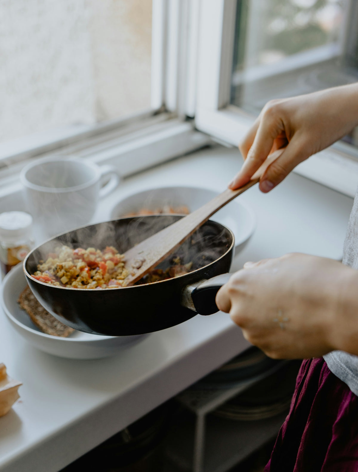 Hands stirring a steaming frying pan with a wooden spatula beside bowls on a sunny windowsill